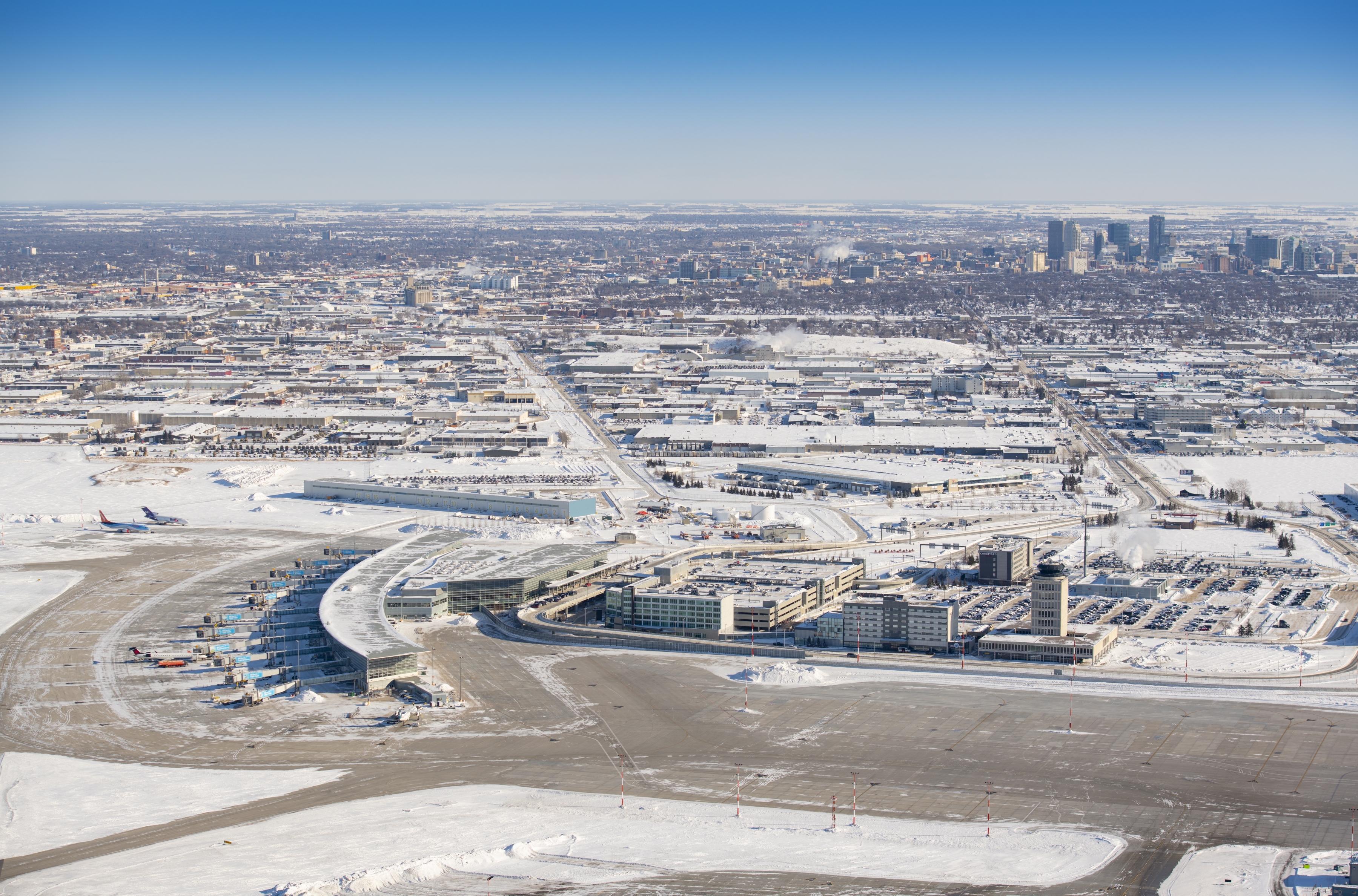 An aerial photo of the Winnipeg Richardson International Airport terminal and surrounding buildings with the city skyline in the background.